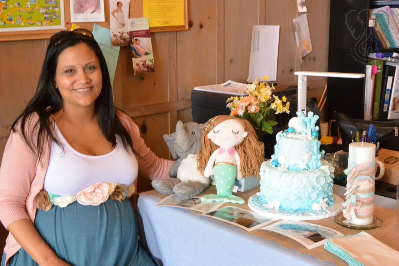 Miranda in front of the ocean-themed cake at Peanut's baby shower. Peanut's mermaid and Adrian's elephant are positioned next to the cake. Miranda is smiling at the camera (photo used with permission)