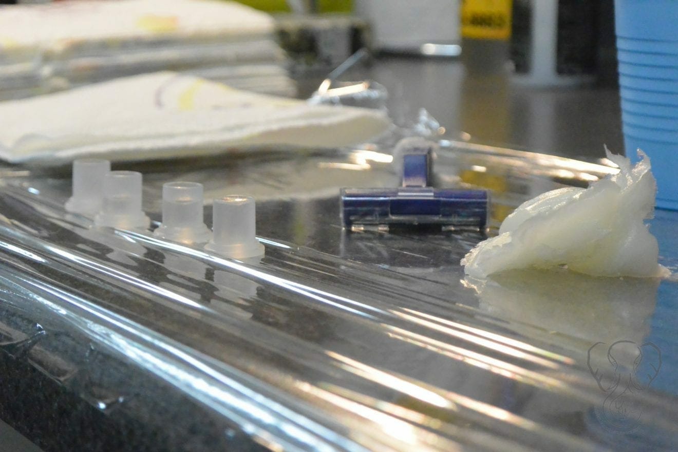 Close up of tattoo preparation table: there are paper towels, ink wells, a razer, and a lob of Vaseline sitting on top of a saran-wrapped table (Miranda Hernandez)